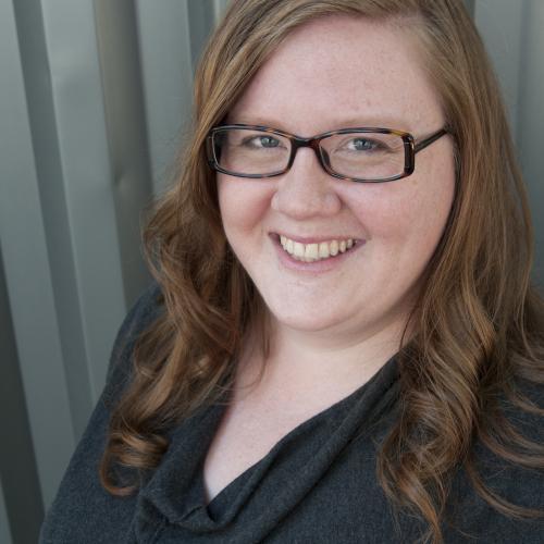 Sabrina has long auburn hair with a light curl and wears tortoise shell glasses and a blue sweater.